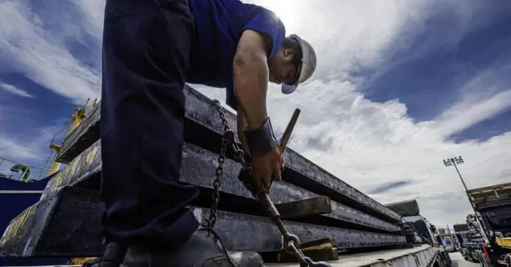 A worker securing long metal cargo on a flatbed trailer using chains to ensure stability during transport. Proper load securing is essential for preventing shifting and ensuring safe delivery.