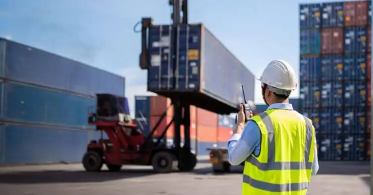 A port worker in a helmet and high-visibility vest holds a walkie-talkie while monitoring the container loading process at a busy port. In the background, a forklift lifts a shipping container, ensuring proper cargo handling and transport efficiency.