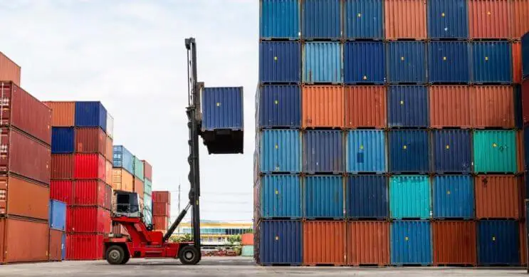 A reach stacker lifting a loaded shipping container onto a stack at a busy port terminal. Efficient container handling with reach stackers helps optimize cargo operations, ensuring safe and balanced container stacking while improving port efficiency.